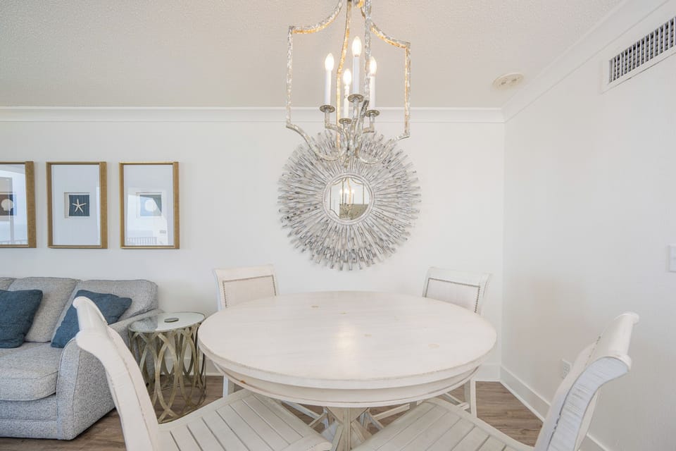 Dining area features a round white table surrounded by stylish chairs, complemented by a decorative mirror and chandelier