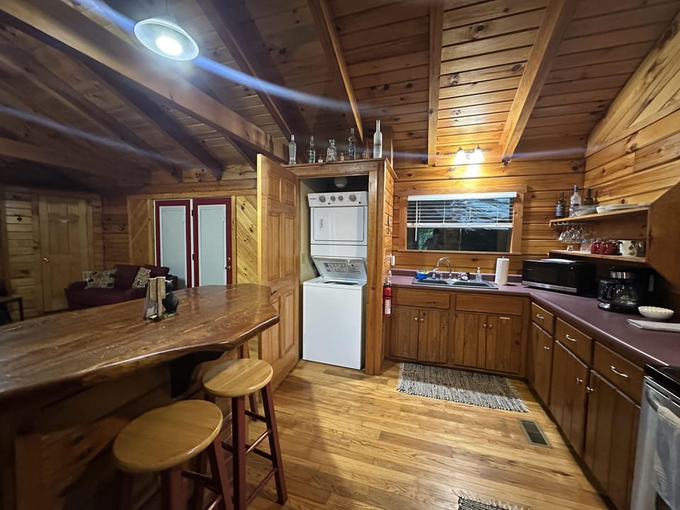 Stacked washer and dryer sit in a wood-paneled nook beside a windowed kitchen sink and purple laminate counters.