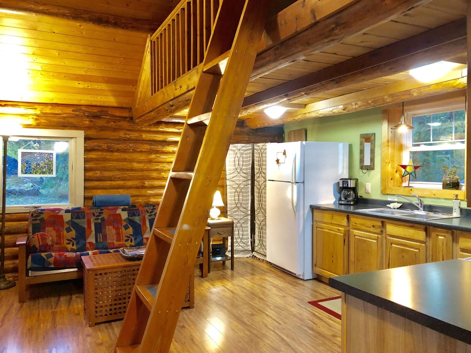 Wooden ladder leads to loft above a kitchen with natural cabinetry, black counters, and a white fridge.