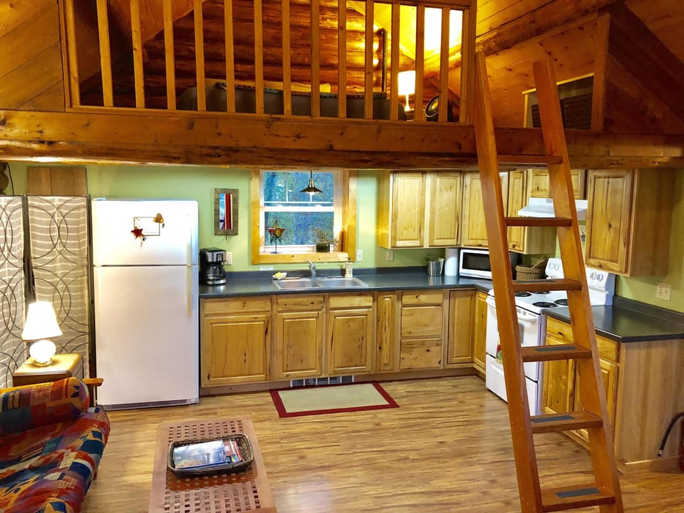 Natural wood cabinetry lines the kitchen walls, with a white fridge, stove, and a ladder ascending to the loft above.