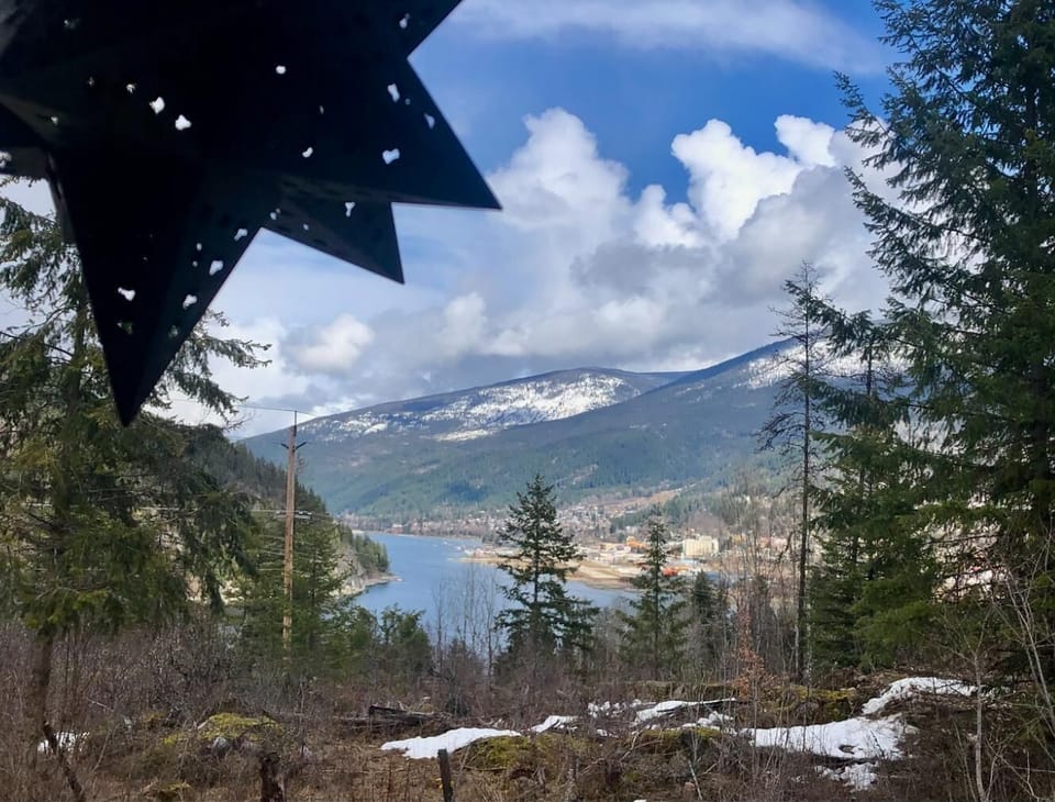 View from cabin window framed by pine trees and black metal star, overlooking snowy mountains and lake.