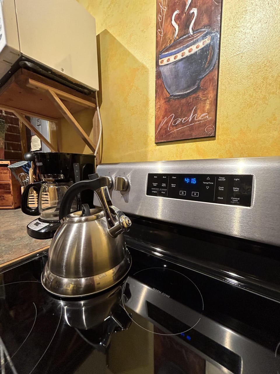 Electric stove with stainless steel kettle reflects on black glass cooktop beneath a mocha-themed wall painting.