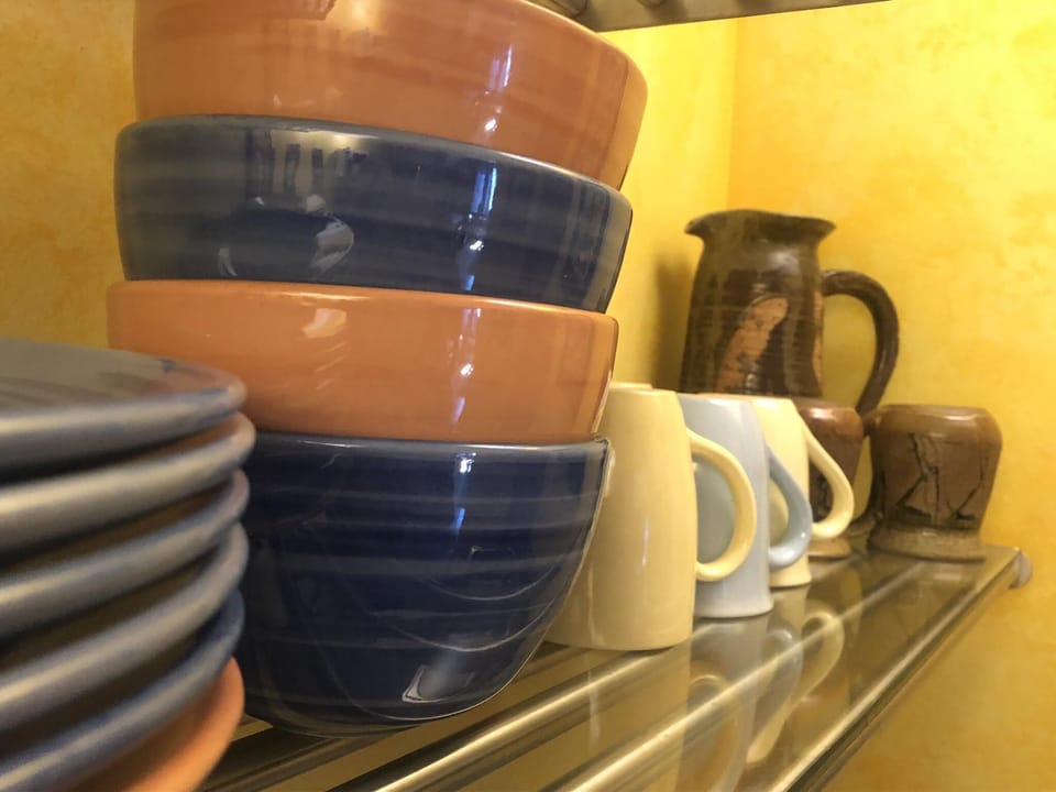 Stacked ceramic bowls and plates sit alongside pastel mugs and rustic pottery on a glass shelf against yellow walls.