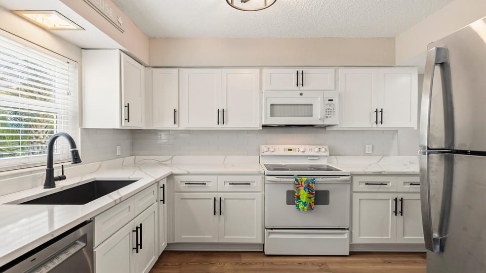 White cabinets with black hardware frame the marble-effect countertops and black sink under a large window.