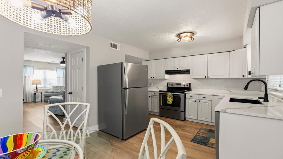 Dining area with glass-top table and white wicker chairs adjacent to kitchen with stainless steel appliances.