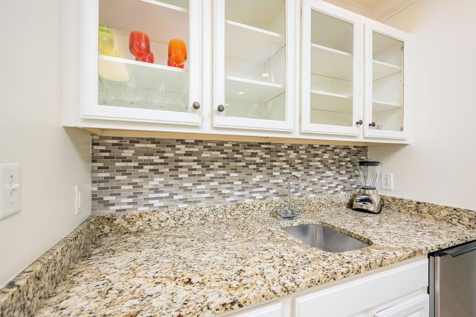 This kitchen features elegant granite countertops, a sleek stainless refrigerator, and a stylish wet bar with glassware