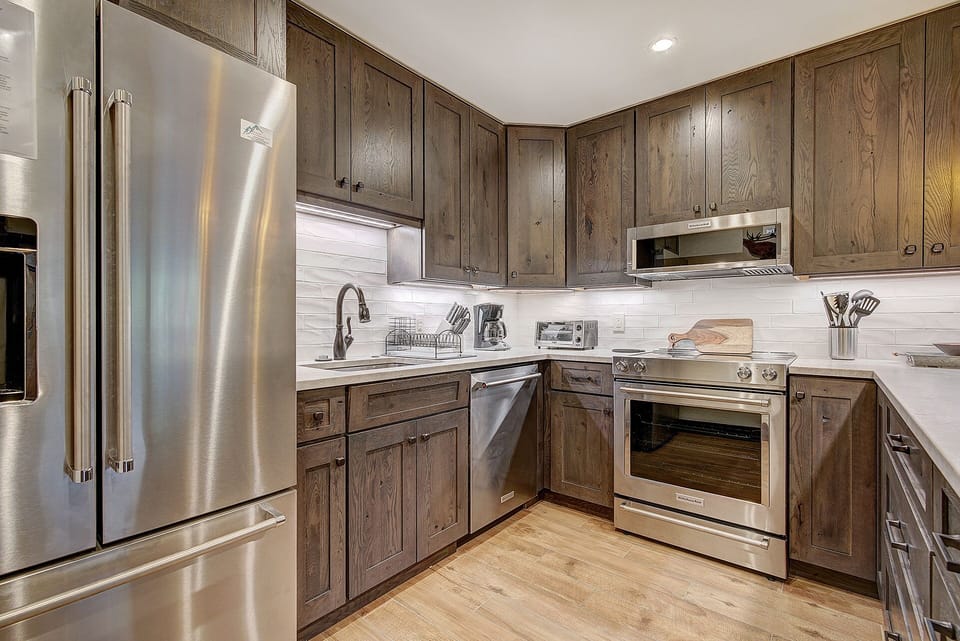 A modern kitchen with stainless steel appliances, dark wood cabinets, a double-door refrigerator, an oven, a microwave, countertop accessories, and a white tile backsplash.
