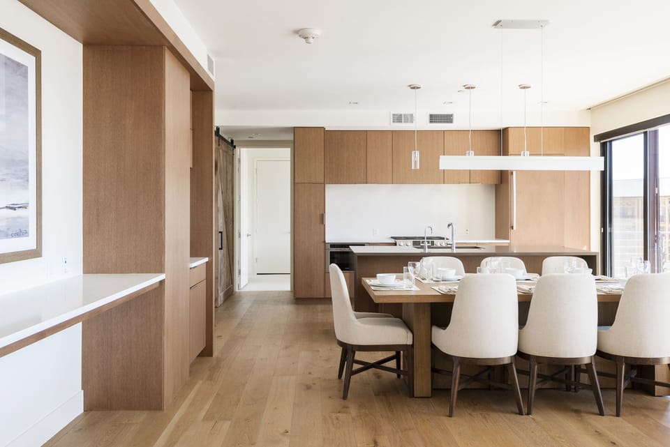 Dining room with linear chandelier light, kitchen in the background and wooden cabinetry