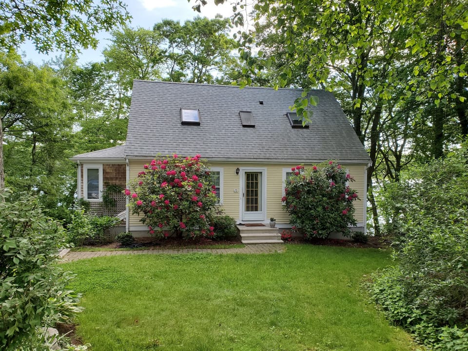 Front view of cottage, which has many flowering bushes, and a vegetable garden.