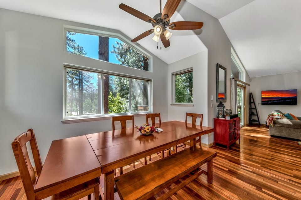Dining area features a wooden table with bench seating, high ceilings, and large windows overlooking pine trees.