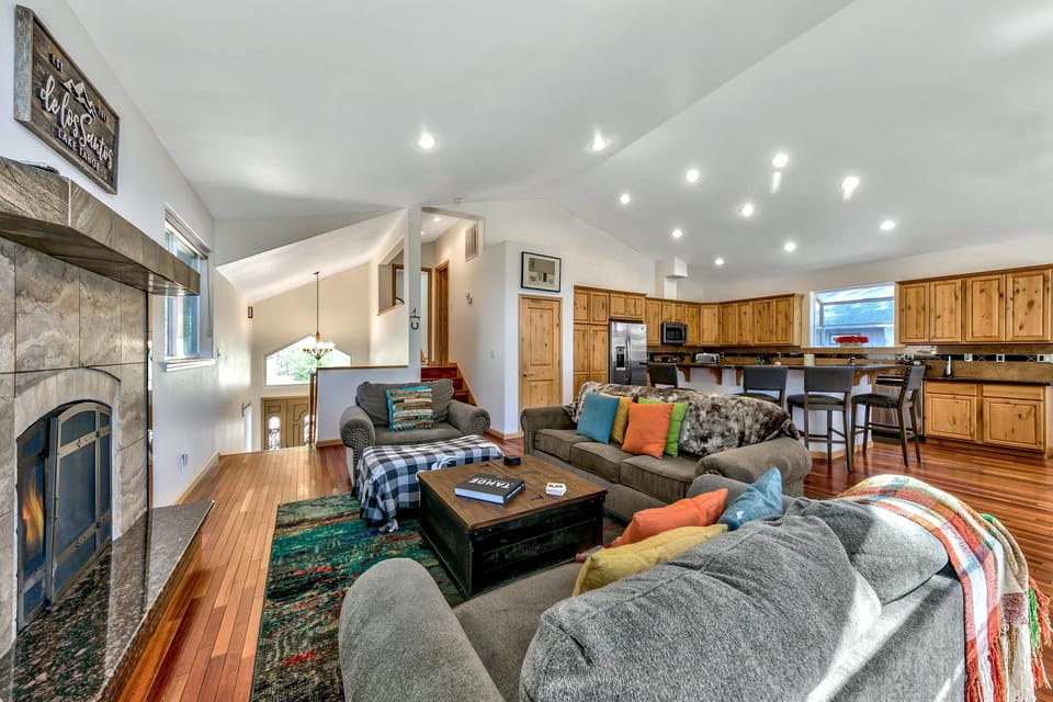 Sunlight brightens the living room with gray upholstery, colorful pillows, a patterned rug, and a stone fireplace.