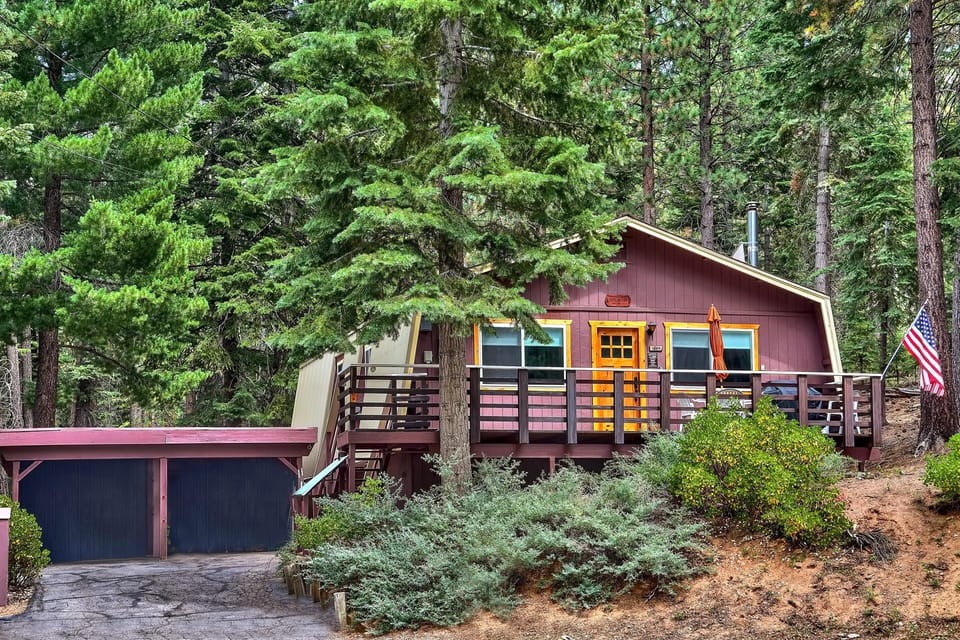 Exterior view of a purple mountain house with wood-trimmed windows, a deck, and attached two-car garage surrounded by pine trees.