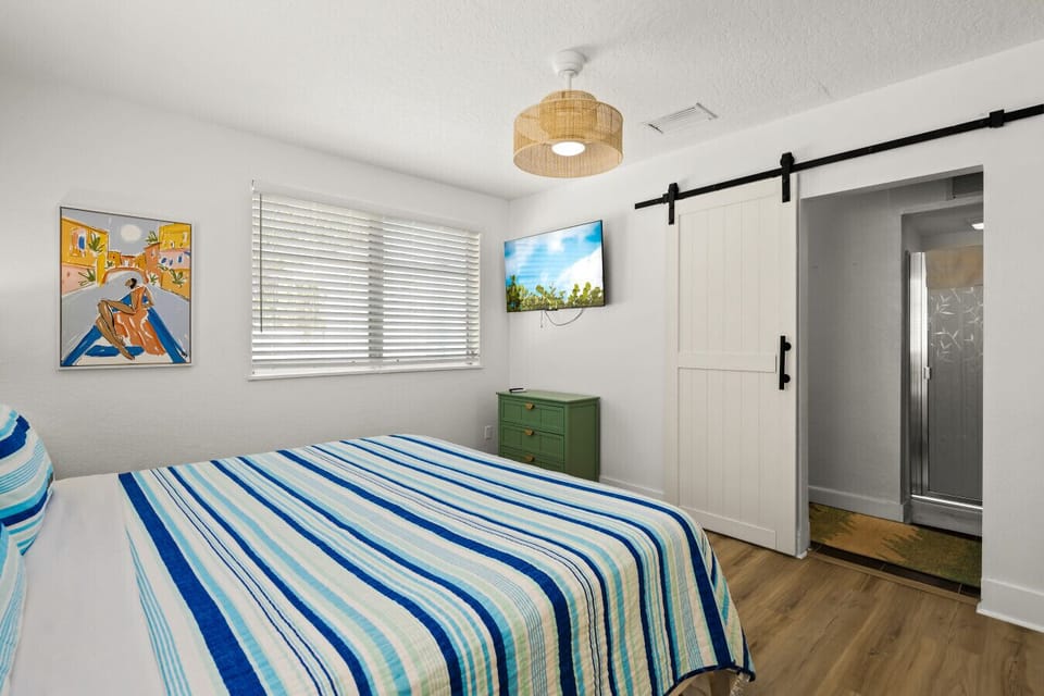 Siesta Key master bedroom with coastal-inspired bedding, seaside art & a barn-door bath—perfect for relaxed coastal living and breezy comfort.
