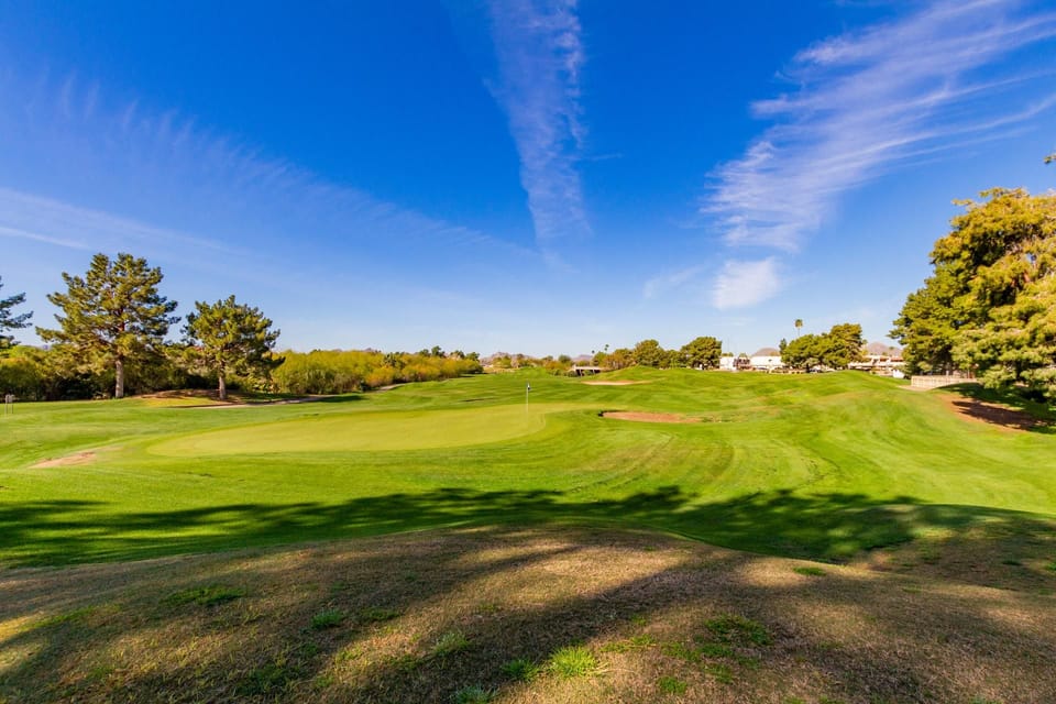Expansive golf course green stretches under a vibrant blue sky, framed by tall trees and distant mountains.