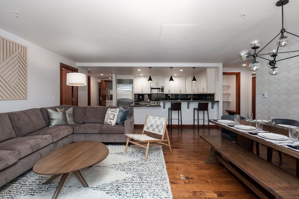 Living room with large area rug, suede sectional, modern wooden coffee table and crosshatch accent chair, dining room with wooden table and modern geometric chandelier
