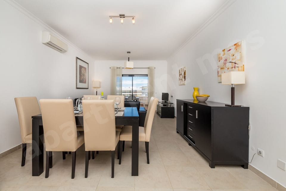 Spacious dining room with six-seater dark wood table, cream upholstered chairs, chandelier and sideboard at Apartment Mos, a professionally cleaned 2-bedroom holiday apartment with communal pool in the Algarve, Portugal