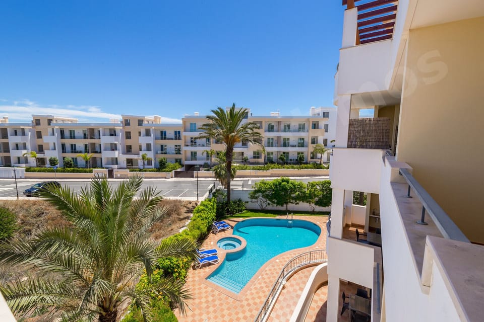 Communal swimming pool surrounded by landscaped gardens, palm trees and the apartment complex with sun loungers on the terrace at Apartment Mos, a professionally cleaned 2-bedroom holiday apartment in the Algarve, Portugal