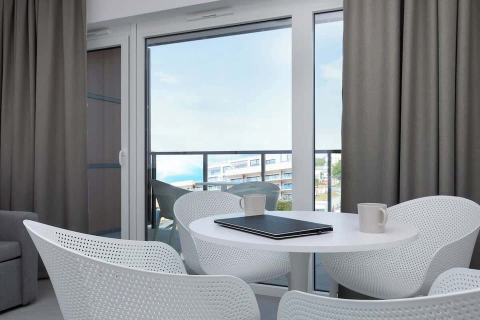 A small dining setup next to large windows, featuring a white table and chairs. The view includes the greenery outside.