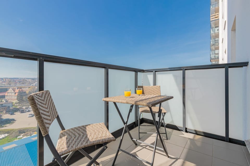 A balcony with a glass railing, featuring a small round table and two wicker chairs. The view overlooks a cityscape with high-rise buildings and greenery.