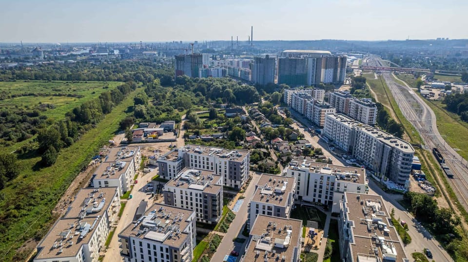 A large aerial view of a modern residential area with several tall apartment buildings. Green spaces and pathways are visible between the buildings.