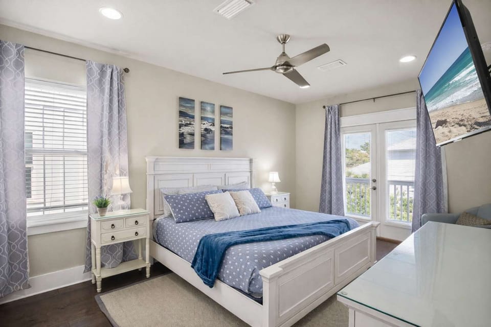 Relax in this spacious primary bedroom featuring a coastal white bed frame, wall-mounted TV, ceiling fan, and French doors leading to a private balcony.