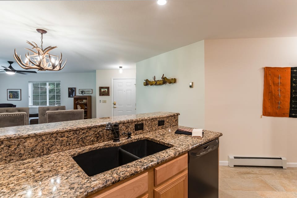 Modern kitchen with granite countertops, a black sink, and wooden cabinets. An antler chandelier hangs over a dining area with a view of a living room and the front door in the background.