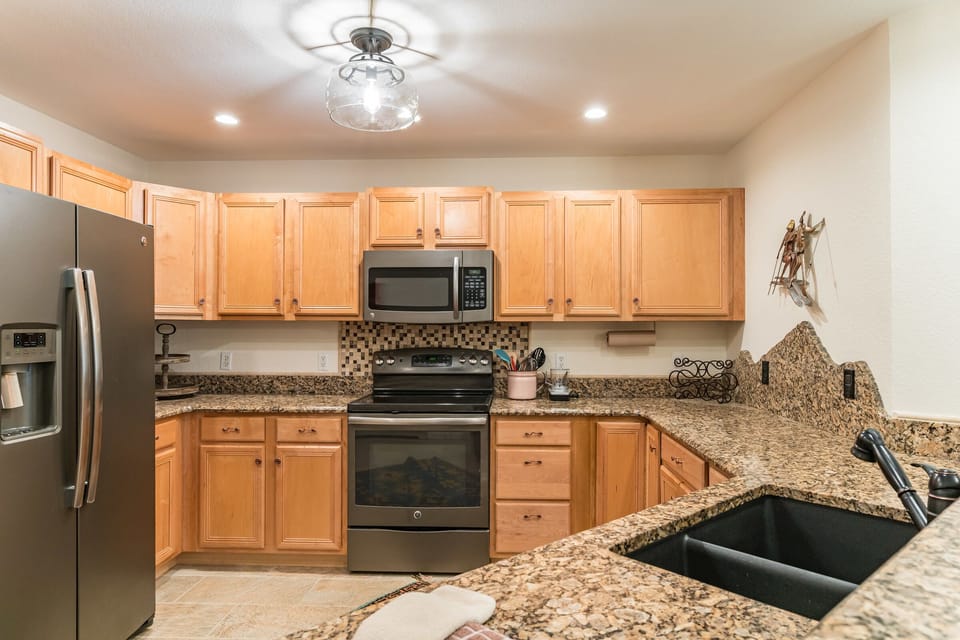 A modern kitchen with light wooden cabinets, a stainless steel refrigerator, an oven with a microwave above, and a granite countertop with a black sink.
