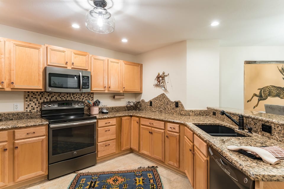 A kitchen with wooden cabinets, granite countertops, a stainless steel oven and microwave, a black sink, and a decorative rug on the floor. Wall decor includes a metal figure and a painting of a bull.