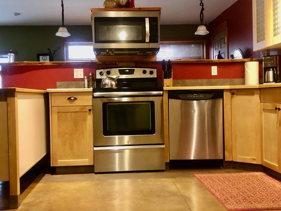 Polished concrete flooring extends beneath natural wood cabinets and a red wall, with a patterned rug near stainless steel appliances.