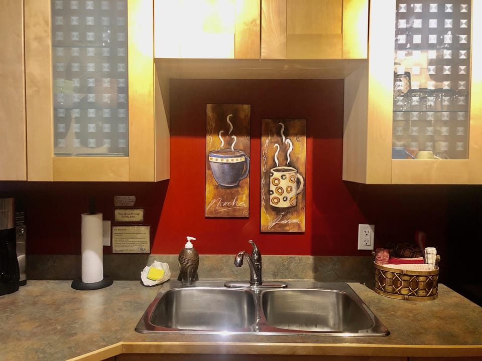 Double stainless steel sink set in stone-look countertop beneath maple cabinets and red accent wall with coffee art.