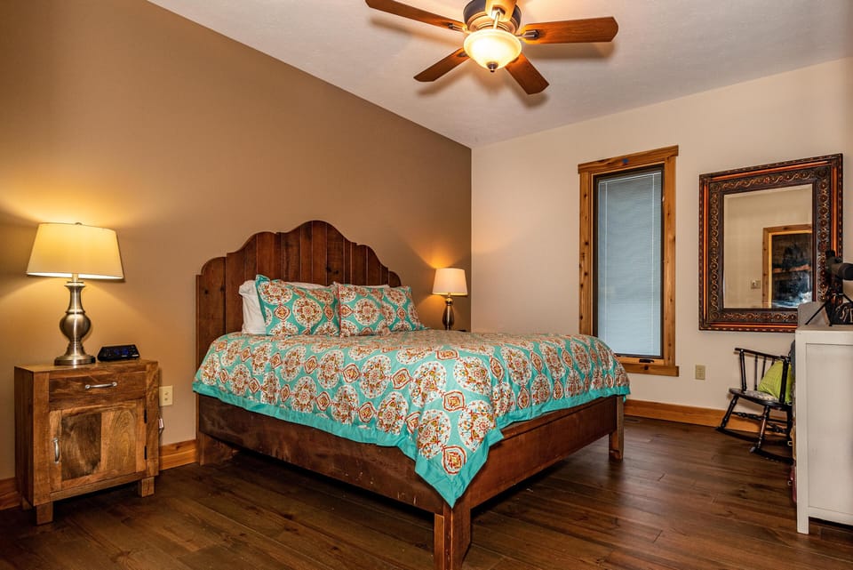 Neutral-toned bedroom with wood trim window, ornate framed mirror, rocking chair, and ceiling fan lighting.