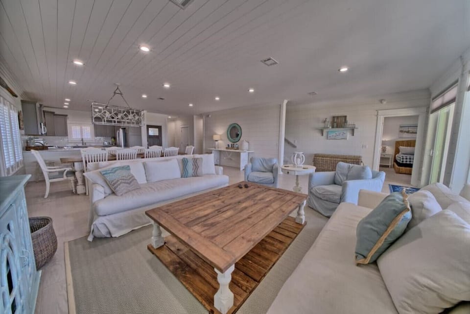 Open concept living room with two white sofas, two armchairs, large wooden coffee table, dining area, and kitchen beyond.
