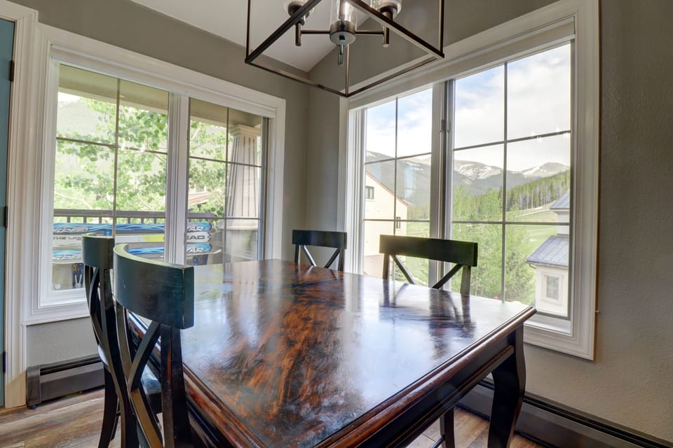 A dining area with a wooden table and four chairs, large windows showcasing a view of mountains and a balcony with trees visible through the glass.