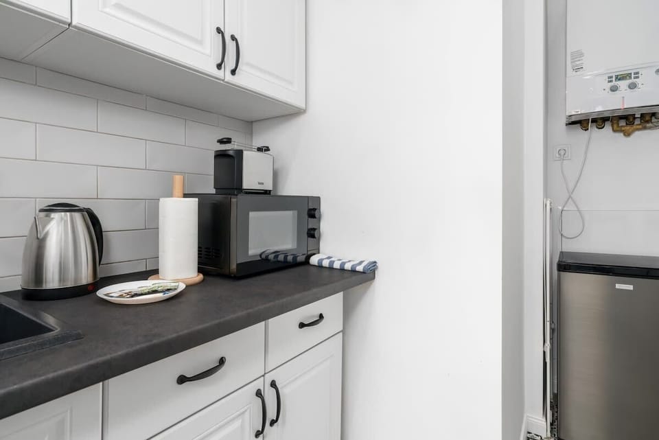 A kitchenette with white cabinetry, a sink, a kettle, and an organized countertop, emphasizing simplicity.