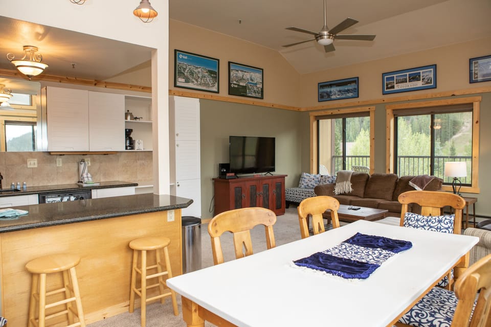 A cozy living space featuring a dining area with a white table and wooden chairs, a kitchen with a counter and stools, and a living room with a couch, TV, and large windows. Framed pictures adorn the walls.