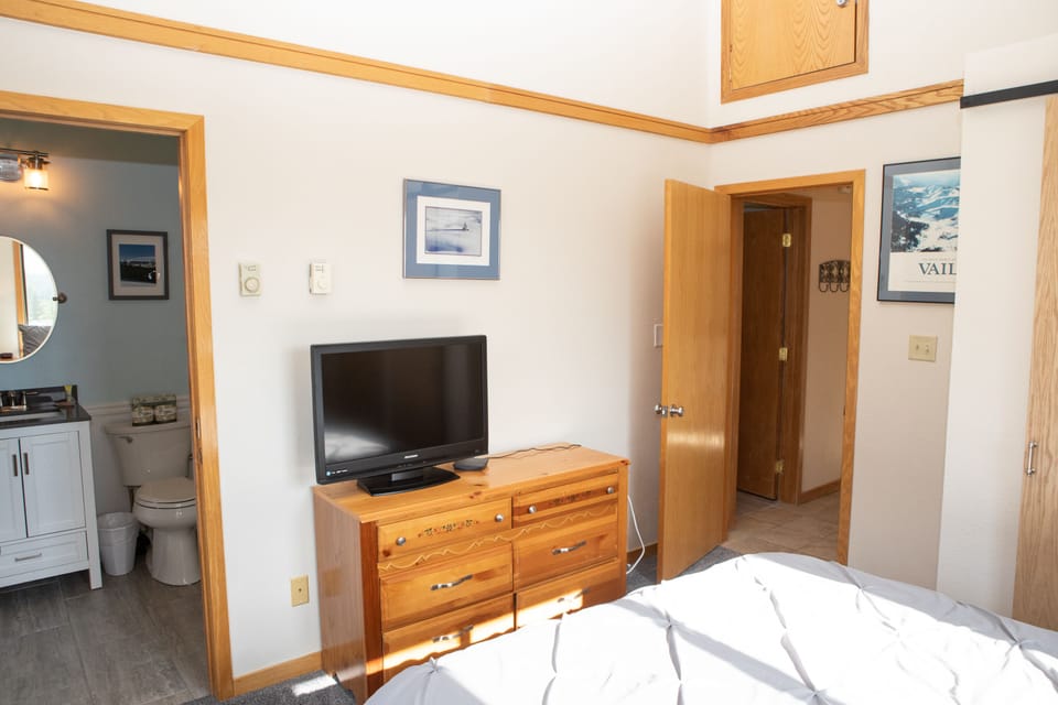 A bedroom with a TV on a wooden dresser, a partially visible bed, and an adjacent bathroom. The walls have framed pictures, and the room features wooden doors and trim.