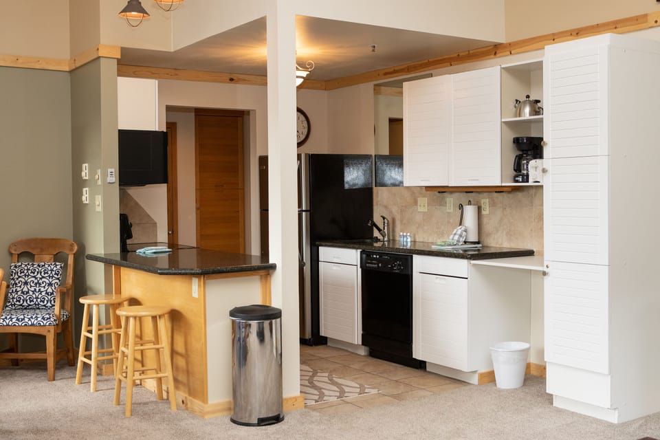 A kitchenette area with white cabinets, black countertops, a small dining area with two stools, a black refrigerator, and a dishwasher. The space includes a small living area with a patterned chair.