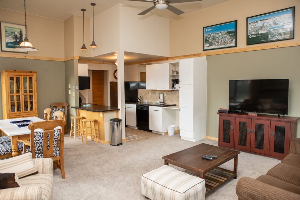 A modern living and dining area with a small kitchen. It includes a table with chairs, TV, sofa, and coffee table. A ceiling fan, posters, and light fixtures are visible. The space has beige and green walls.