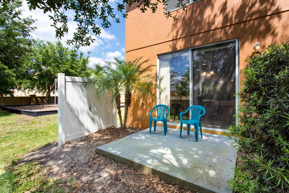 Concrete patio with two turquoise plastic chairs, sliding glass door, and nearby palm trees and white privacy fence.