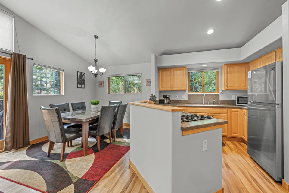 Spacious open-concept dining area with elegant chandelier lighting and a fully equipped kitchen featuring natural wood cabinetry and abundant counter space. Large windows provide plenty of natural light and a tranquil wooded view.