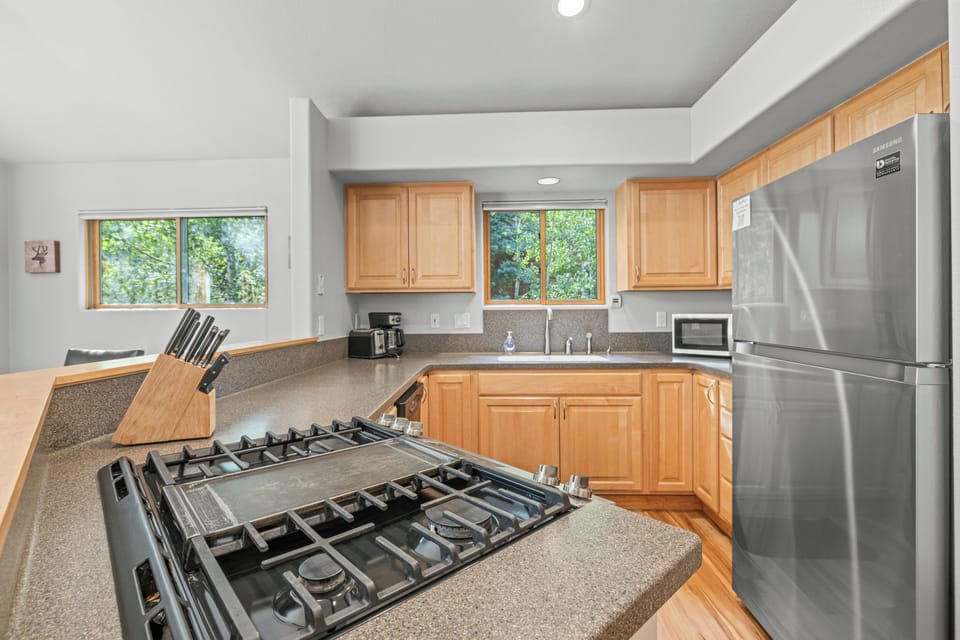 Spacious kitchen boasting natural wood cabinetry, a stainless steel refrigerator, and a sleek gas cooktop. Sunlight streams through large windows, creating a warm and inviting space to cook and gather after a day exploring the mountains.