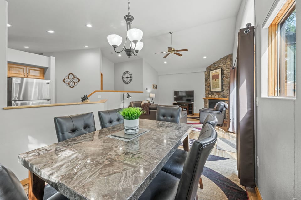 Stylish dining room featuring a polished stone-top table and plush seating for six. Large windows and glass doors frame a peaceful forest backdrop, seamlessly connecting to the cozy living area and stone fireplace.