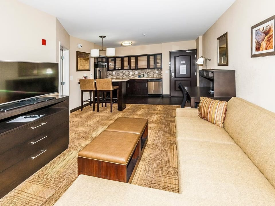 Living area with sofa, ottoman, and work desk — kitchen with bar stools visible in the background