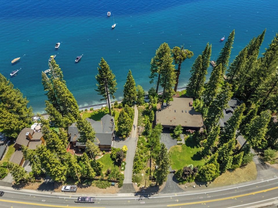 Aerial view of a lakeside property nestled among tall pines, with crystal-clear blue waters, private beach access, and boats moored just offshore.