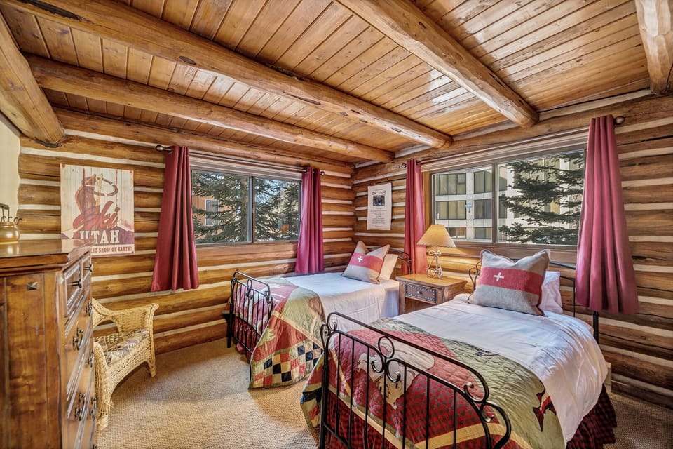 This bedroom is furnished with one king bed or two twin beds and has colorful quilted bedding, red curtains, wood walls, and windows overlooking the surrounding buildings.