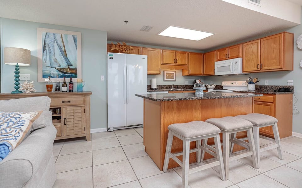 Cozy kitchen area features light wood cabinetry, a breakfast bar with three stools, and a decorative coastal-themed artwork