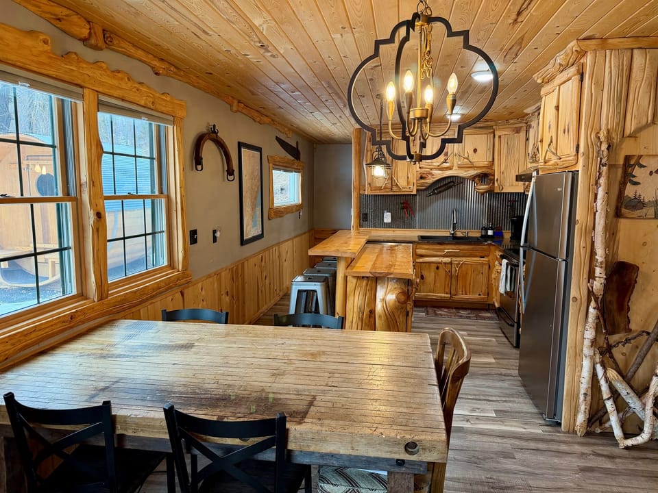 A warm, modern rustic kitchen blends natural wood tones with sleek stainless steel. The farmhouse style dining table anchors the space, creating a cozy yet stylish gathering spot that feels both cabin-inspired and beautifully updated.