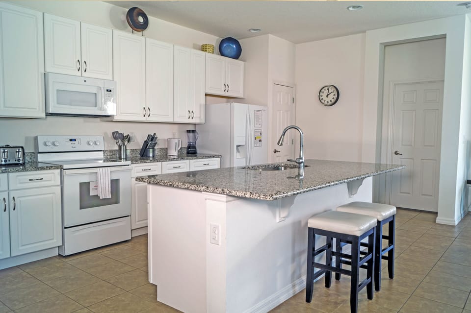 Open white kitchen with large granite island and two barstools.