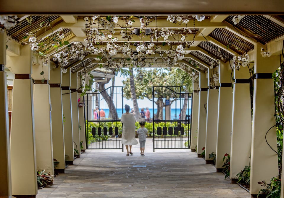A charming pathway framed by flowering vines leads to the property's beachfront entrance, with ocean views and tropical surroundings beyond the gates.