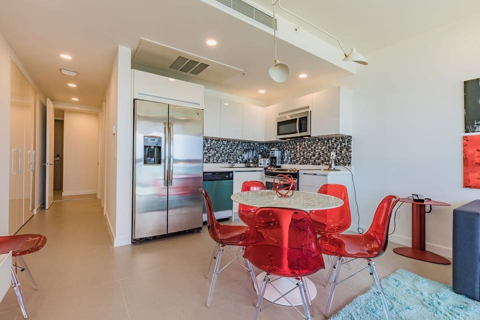 Your modern open-concept kitchen features stainless steel appliances, sleek white cabinetry, and a stylish mosaic backsplash. The bright dining space with bold red chairs invites you to gather and enjoy meals in contemporary comfort.